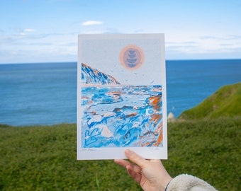 Puede incluir: Una impresión azul y naranja de un paisaje marítimo con un acantilado y un sol con tres cuencos apilados. La impresión se sostiene frente a un fondo borroso de una colina verde cubierta de hierba y un océano azul.