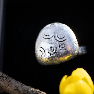 Könnte beinhalten: Ein silberner Ring mit einem einzigartigen Design aus wirbelnden Mustern. Der Ring ist oval geformt und hat eine glatte, polierte Oberfläche.