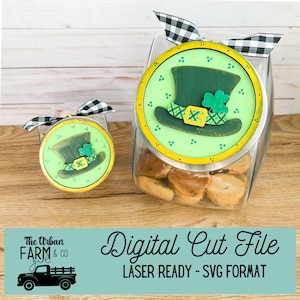 Può includere: Due barattoli di biscotti di vetro con coperchi decorati con cappelli da folletto verdi. I coperchi sono rotondi e hanno un bordo giallo. I barattoli sono pieni di biscotti. Il testo "The Urban Farm Girl & Co." è su uno sfondo blu con una grafica di camion nero. Il testo "Digital Cut File" è in un carattere bianco corsivo su uno sfondo blu. Il testo "LASER READY - SVG FORMAT" è in testo nero su uno sfondo blu.