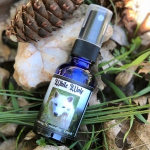 May include: A blue glass bottle with a white wolf image and the text "White Wolf" and "Calming * Grounding * Empower" on the label. The bottle has a black spray nozzle and is sitting on a bed of pine needles and green grass.