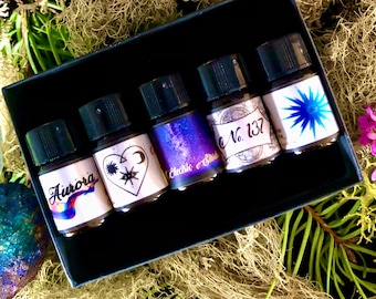 May include: A set of five small glass bottles with black caps and colorful labels. The labels feature celestial designs and text, including "Aurora", "Electric Bliss", and "No. 137". The bottles are arranged in a black box.