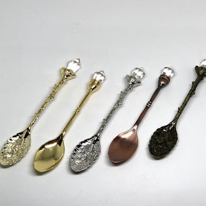 May include: Five ornate spoons with clear crystal handles. The spoons are made of different metals, including gold, silver, and copper. Each spoon has a unique design on the bowl.