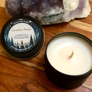 May include: A black metal tin candle with a white label that reads "Coconut Soy Candle Lavender & Amethyst. FOREST & FOLKLORE Net Wt. 4 oz. (114 g) www.forestandfolklore.com". The candle is lit and the wax is melting.