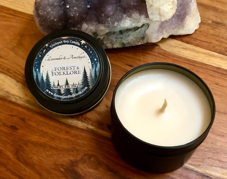 May include: A black metal tin candle with a white label that reads "Coconut Soy Candle Lavender & Amethyst. FOREST & FOLKLORE Net Wt. 4 oz. (114 g) www.forestandfolklore.com". The candle is lit and the wax is melting.