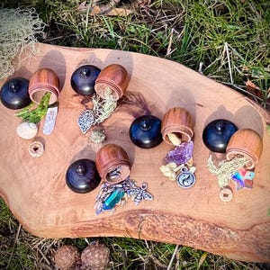 May include: Six wooden acorn-shaped boxes with lids open, revealing various small trinkets and crystals. The boxes are arranged on a wooden surface with a green grassy background.