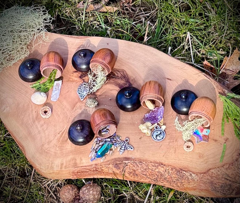 May include: Six wooden acorn-shaped boxes with lids open, revealing various small trinkets and crystals. The boxes are arranged on a wooden surface with a green grassy background.