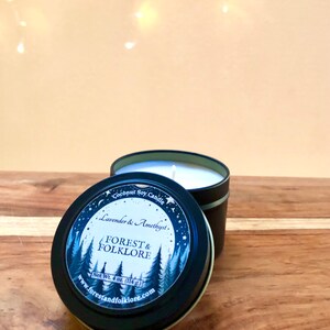 May include: A black tin candle with a white label. The label features a forest scene with trees and a full moon. The text on the label reads "Coconut Soy Candle", "Lavender & Amethyst", "Forest & Folklore", "Net Wt. 4 oz. (114 g.)", and "www.forestandfolklore.com".