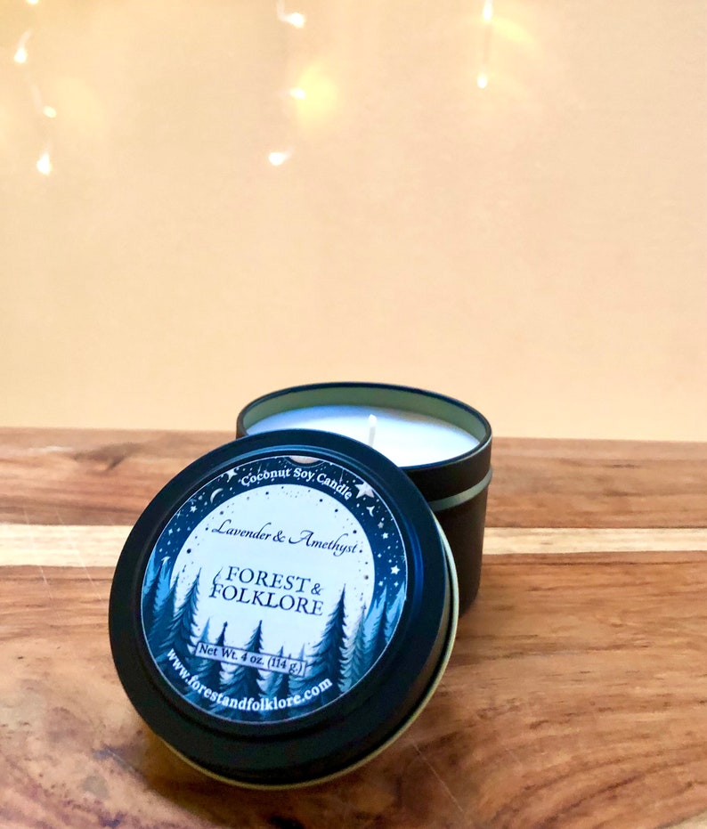 May include: A black tin candle with a white label. The label features a forest scene with trees and a full moon. The text on the label reads "Coconut Soy Candle", "Lavender & Amethyst", "Forest & Folklore", "Net Wt. 4 oz. (114 g.)", and "www.forestandfolklore.com".
