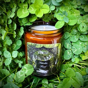 May include: A brown glass jar candle with a white wick, labeled "Forest & Folklore - Pohickor Forest." The candle is surrounded by vibrant green clover leaves, creating a natural, earthy aesthetic. The label features a forest scene.