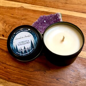 May include: A black tin candle with a white label that reads "Coconut Soy Candle Lavender & Amethyst. FOREST & FOLKLORE Net Wt. 4 oz (114g) www.forestandfolklore.com". The candle is sitting on a wooden surface next to a purple amethyst crystal.