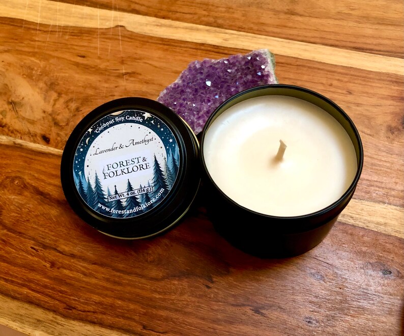 May include: A black tin candle with a white label that reads "Coconut Soy Candle Lavender & Amethyst. FOREST & FOLKLORE Net Wt. 4 oz (114g) www.forestandfolklore.com". The candle is sitting on a wooden surface next to a purple amethyst crystal.
