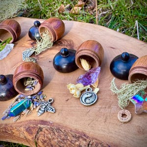 May include: Six wooden acorn-shaped containers with lids, each filled with different crystals, herbs, and other small objects. The containers are arranged on a wooden surface with a green background.