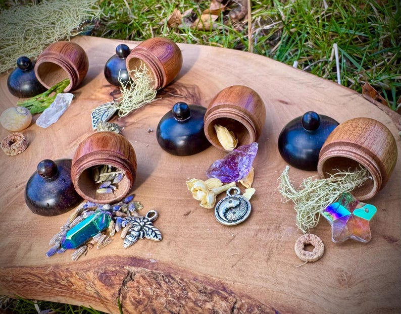 May include: Six wooden acorn-shaped containers with lids, each filled with different crystals, herbs, and other small objects. The containers are arranged on a wooden surface with a green background.