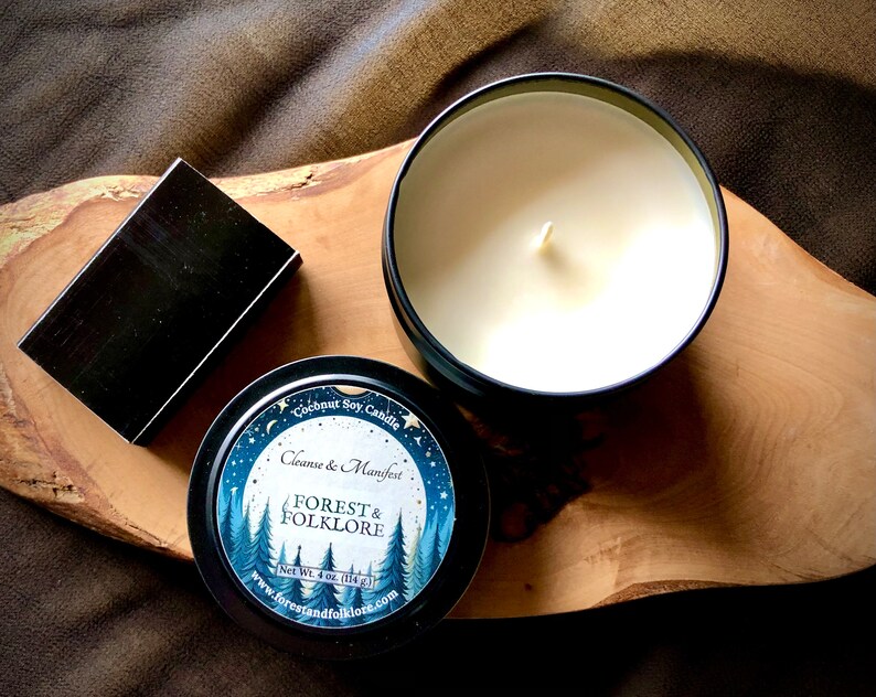May include: A black tin candle with a white label that says "Coconut Soy Candle Cleanse & Manifest FOREST & FOLKLORE Net Wt. 4 oz. (114g) www.forestandfolklore.com". The label has a blue and white illustration of a forest with a full moon and stars.