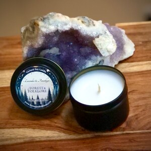 May include: A black metal tin candle with a white label that reads "Coconut Soy Candle, Lavender & Amethyst, Forest & Folklore, Handmade with Love, www.forestandfolklore.com". The candle is sitting on a wooden surface next to a large amethyst geode.