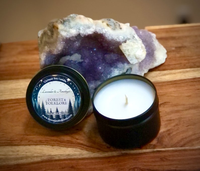 May include: A black metal tin candle with a white label that reads "Coconut Soy Candle, Lavender & Amethyst, Forest & Folklore, Handmade with Love, www.forestandfolklore.com". The candle is sitting on a wooden surface next to a large amethyst geode.