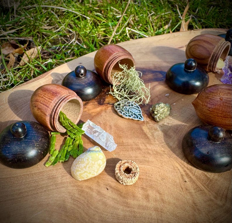 May include: A collection of small wooden acorn-shaped boxes with lids, some open to reveal green moss, green plant material, and a clear crystal. The boxes are arranged on a wooden surface with other small objects, including a silver leaf, a small rock, and a ring.