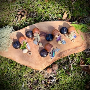 May include: Seven small wooden acorn-shaped boxes with lids open, revealing various small trinkets and crystals inside. The boxes are arranged on a wooden surface with a green grassy background.
