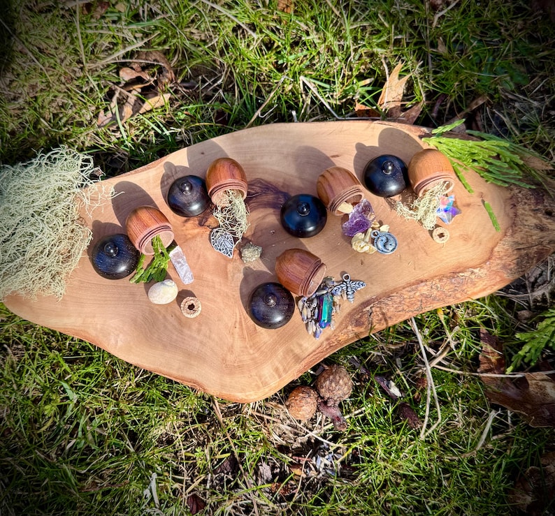 May include: Seven small wooden acorn-shaped boxes with lids open, revealing various small trinkets and crystals inside. The boxes are arranged on a wooden surface with a green grassy background.