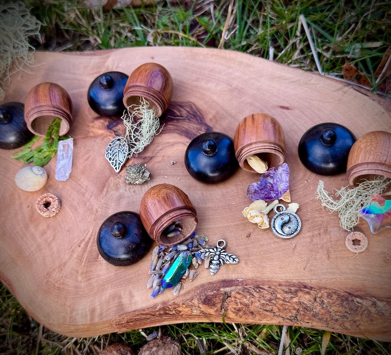 May include: A collection of small wooden acorn-shaped boxes with black lids, open to reveal various crystals, dried flowers, and other small trinkets. The boxes are arranged on a wooden surface with green foliage in the background.