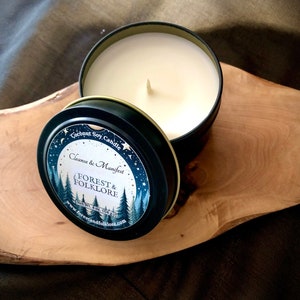 May include: A black tin candle with a white label that reads "Coconut Soy Candle Cleanse & Manifest FOREST & FOLKLORE Net Wt. 4 oz (114 g) www.forestandfolklore.com". The candle is on a wooden surface.