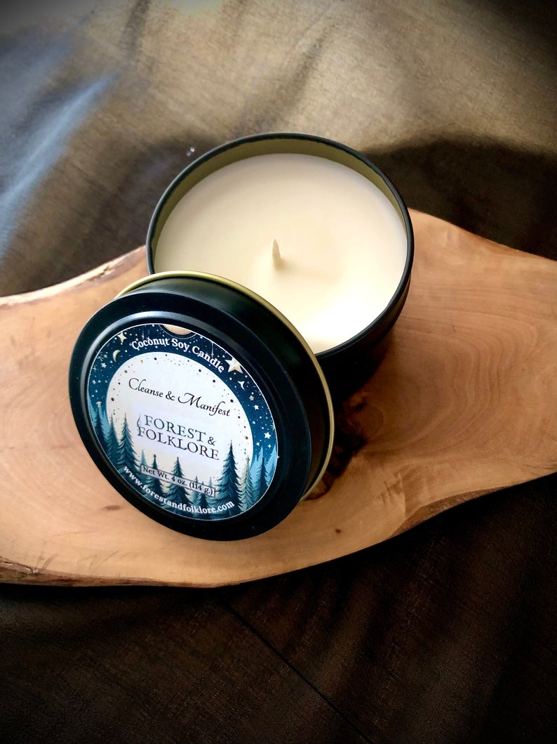 May include: A black tin candle with a white label that reads "Coconut Soy Candle Cleanse & Manifest FOREST & FOLKLORE Net Wt. 4 oz (114 g) www.forestandfolklore.com". The candle is on a wooden surface.