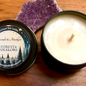 May include: A black tin candle with a white label that reads "Coconut Soy Candle Lavender & Amethyst. FOREST & FOLKLORE Net Wt. 4 oz. (114 g) www.forestandfolklore.com". The candle is sitting on a wooden surface next to a purple amethyst crystal.