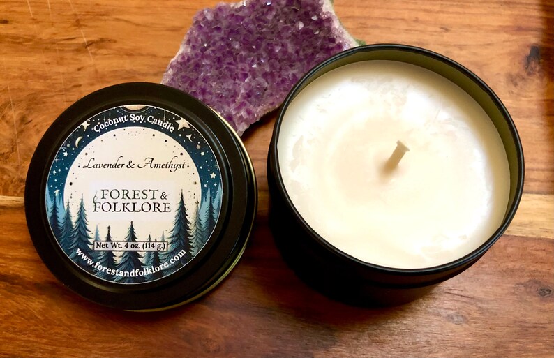 May include: A black tin candle with a white label that reads "Coconut Soy Candle Lavender & Amethyst. FOREST & FOLKLORE Net Wt. 4 oz. (114 g) www.forestandfolklore.com". The candle is sitting on a wooden surface next to a purple amethyst crystal.