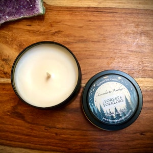 May include: A black candle tin with a white soy candle inside. The lid of the tin has a design of a moon, stars, and trees with the text "Coconut Soy Candle", "Lavender & Amethyst", "Forest & Folklore", "Net Wt. 4 oz (113g)", and "www.forestandfolklore.com".