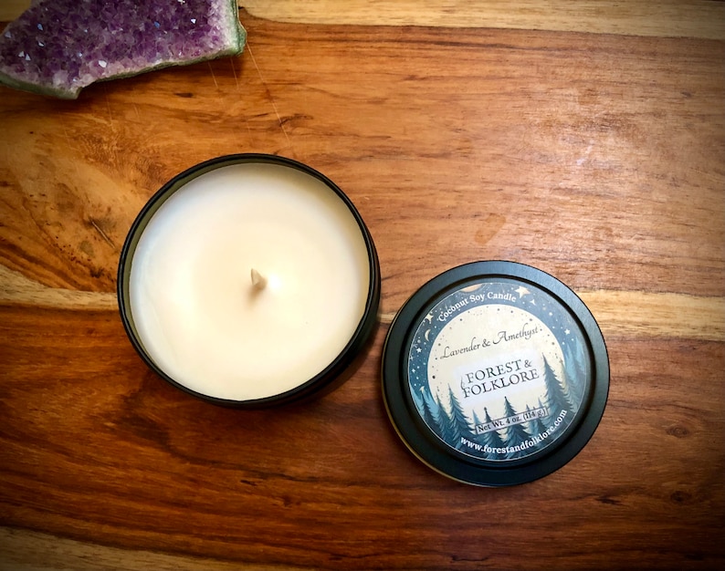 May include: A black candle tin with a white soy candle inside. The lid of the tin has a design of a moon, stars, and trees with the text "Coconut Soy Candle", "Lavender & Amethyst", "Forest & Folklore", "Net Wt. 4 oz (113g)", and "www.forestandfolklore.com".