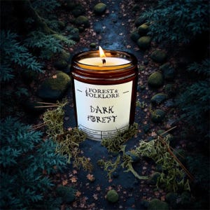 May include: A lit candle in a brown glass jar with a white label that reads "FOREST & FOLKLORE DARK FOREST". The candle is surrounded by moss and foliage, creating a natural, earthy aesthetic. The flame glows brightly against the dark background.