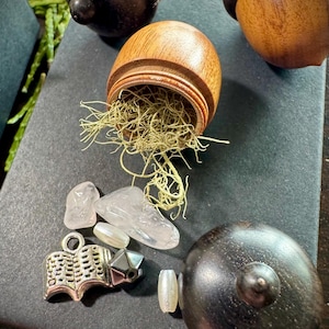 May include: Three wooden acorn-shaped boxes, one open to reveal green moss and clear quartz crystals. A silver charm in the shape of an open book is also visible.