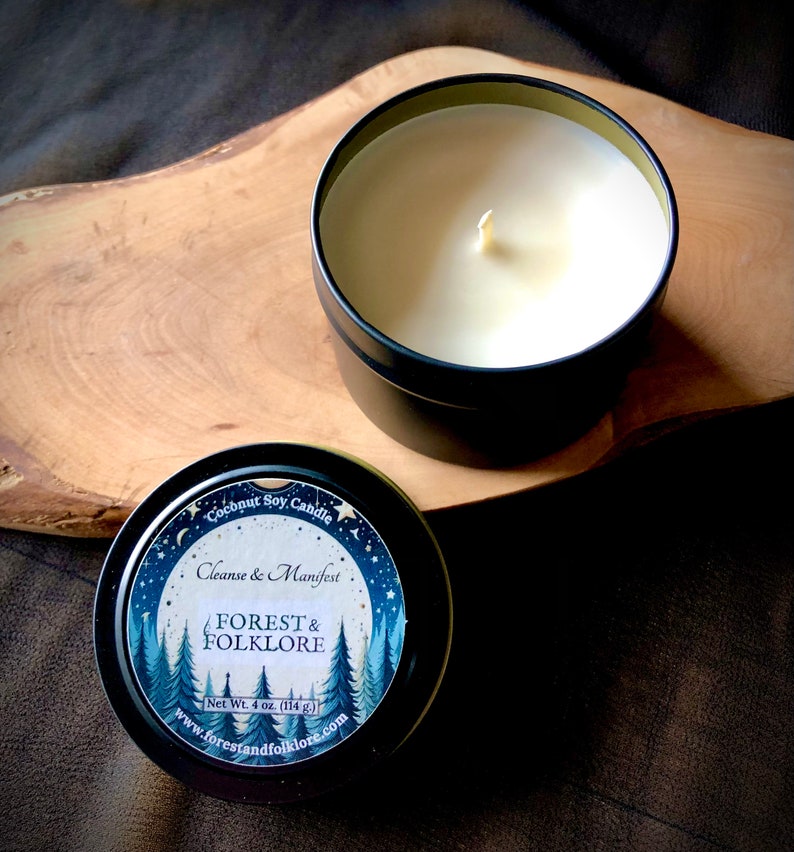 May include: A black metal tin candle with a white label that reads "Coconut Soy Candle, Cleanse & Manifest, FOREST & FOLKLORE, Net Wt. 4 oz. (114 g.), www.forestandfolklore.com". The label features a watercolor illustration of a forest with stars and moons.