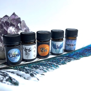 May include: Five small amber glass bottles with black screw-on caps, each featuring a label with the text Earth, Air, Fire, Water, or Spirit. The bottles are arranged in a row alongside a large purple amethyst cluster and a long peacock feather.