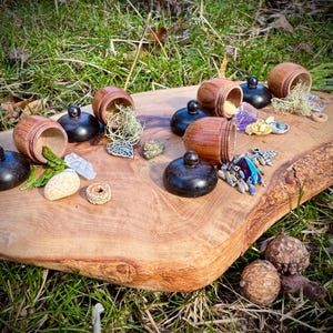May include: A wooden tray with six small wooden acorn-shaped boxes with lids. The boxes are open and contain various small items, including crystals, beads, and a small metal charm. The tray is on a bed of green grass.