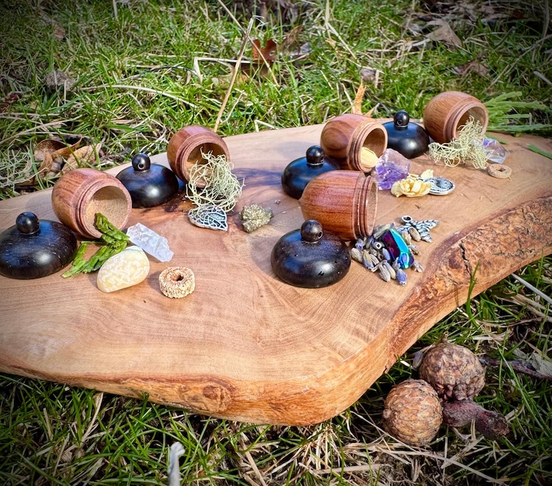 May include: A wooden tray with six small wooden acorn-shaped boxes with lids. The boxes are open and contain various small items, including crystals, beads, and a small metal charm. The tray is on a bed of green grass.