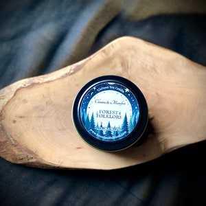 May include: A small, round, black tin candle with a blue and white label. The label reads "Coconut Soy Candle", "Cleanse & Manifest", "Forest & Folklore", "Net Wt. 4 oz (113 g)", and "www.forestandfolklore.com".