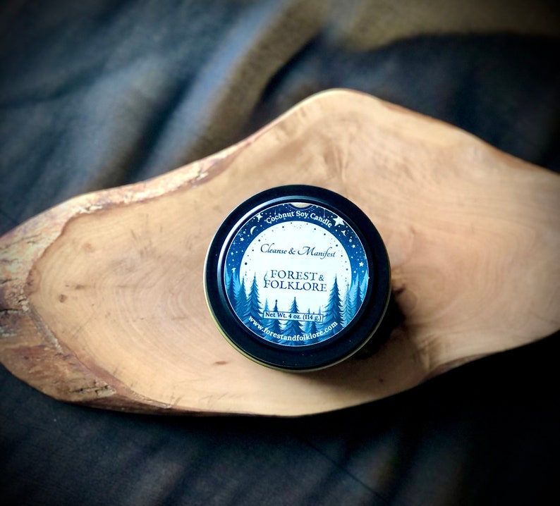 May include: A small, round, black tin candle with a blue and white label. The label reads "Coconut Soy Candle", "Cleanse & Manifest", "Forest & Folklore", "Net Wt. 4 oz (113 g)", and "www.forestandfolklore.com".