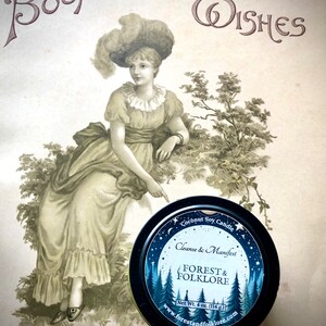 May include: A black tin candle with a blue and white label. The label features a moon, stars, and pine trees. The text on the label reads "Coconut Soy Candle", "Cleanse & Manifest", "Forest & Folklore", "Net Wt. 4 oz. (114 g)", and "www.forestandfolklore.com".