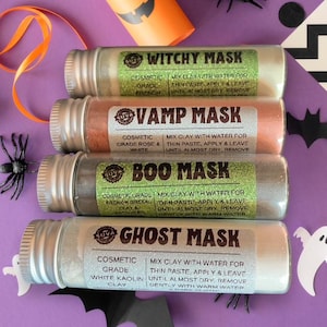 Könnte beinhalten: Vier Glasröhrchen mit kosmetischen Gesichtsmasken mit den Aufschriften "Witchy Mask", "Vamp Mask", "Boo Mask" und "Ghost Mask". Jedes Röhrchen hat einen silbernen Deckel und ein Etikett mit Anweisungen. Die Masken sind in Grün-, Orange- und Weißtönen gehalten.