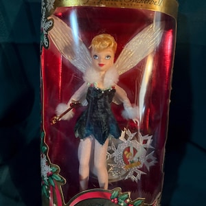 May include: A Holiday Sparkle Tinkerbell Special Edition doll in a clear plastic display case. The doll is wearing a dark green dress with white fur trim and has blonde hair and large white wings. The packaging features the Walt Disney's Peter Pan logo.