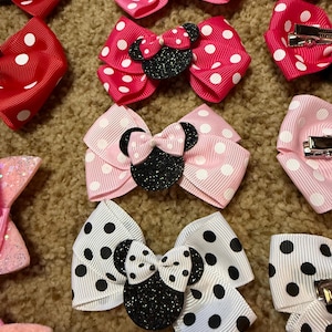 May include: A collection of polka dot hair bows in various colors, including red, pink, and white. Each bow features a glittery black mouse-ear design. The bows are arranged on a textured surface, showcasing their different styles.