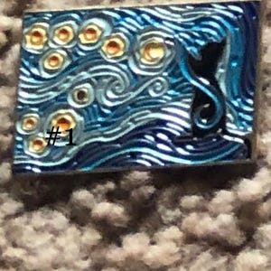 May include: A blue and silver enamel pin with a black silhouette of a cat in front of a swirling, starry night sky. The pin has a number "#1" in the lower left corner.