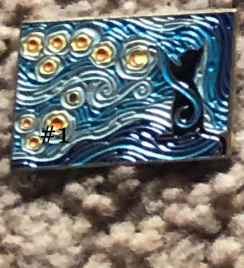 May include: A blue and silver enamel pin with a black silhouette of a cat in front of a swirling, starry night sky. The pin has a number "#1" in the lower left corner.
