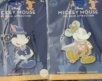 May include: Two Disney Mickey Mouse limited edition pins. The first pin features Mickey Mouse dressed as Peter Pan with a blue and white outfit and a hat. The second pin features Mickey Mouse dressed as Prince Charming with a gold crown and a white and pink outfit. Both pins have the text "Mickey Mouse The Main Attraction" and "Limited Release Edition Limitée".