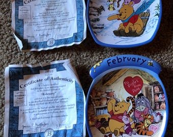May include: Two Winnie the Pooh themed ceramic plates, one for January and one for February. The January plate features Winnie the Pooh on a sled in a snowy scene. The February plate features Winnie the Pooh and Piglet in a kitchen scene with a heart that says "To Piglet from Pooh".