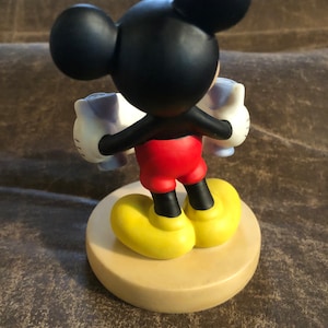Puede incluir: Una figurita de cer&aacute;mica de Mickey Mouse sosteniendo dos tazas blancas con detalles azules. Mickey lleva pantalones cortos rojos, zapatos negros con suelas amarillas y sus guantes negros caracter&iacute;sticos. La figurita est&aacute; de pie sobre una base de color beige.