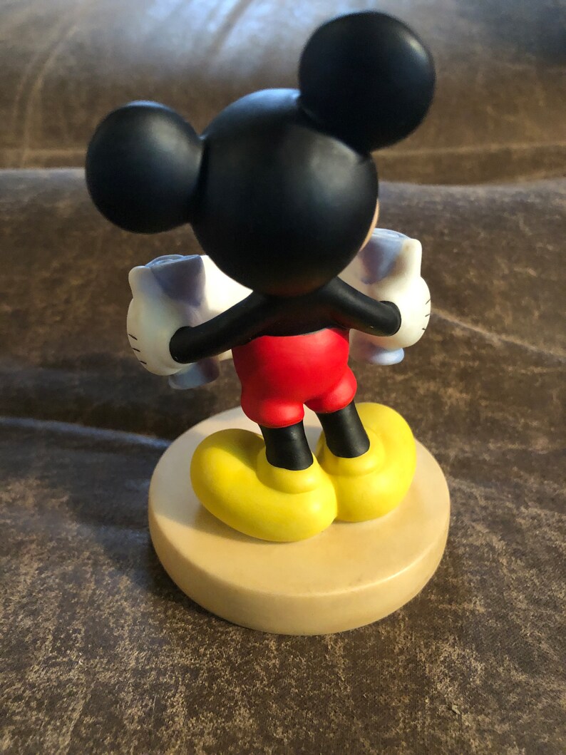 Puede incluir: Una figurita de cer&aacute;mica de Mickey Mouse sosteniendo dos tazas blancas con detalles azules. Mickey lleva pantalones cortos rojos, zapatos negros con suelas amarillas y sus guantes negros caracter&iacute;sticos. La figurita est&aacute; de pie sobre una base de color beige.