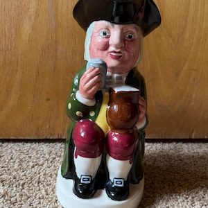 May include: A ceramic figurine of a man in 18th-century attire, holding a mug and a small cup. The figure wears a black hat, green coat, and white wig. The figurine has a white base and is painted with vibrant colors.