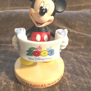Puede incluir: Una figurita de cer&aacute;mica de Mickey Mouse sosteniendo un rollo con el texto "2003 Walt Disney World. The Magical Place To Be." El rollo es blanco con detalles coloridos y la base es beige.